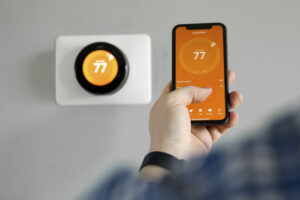 Person uses a mobile phone with smart home app in modern living room, with smart thermostat in wall in background. Both show temp setting of 77 degrees.